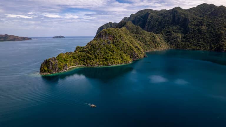 Drone shot of an island full of trees and a deep blue water with one small traditional boat in Philippines.
