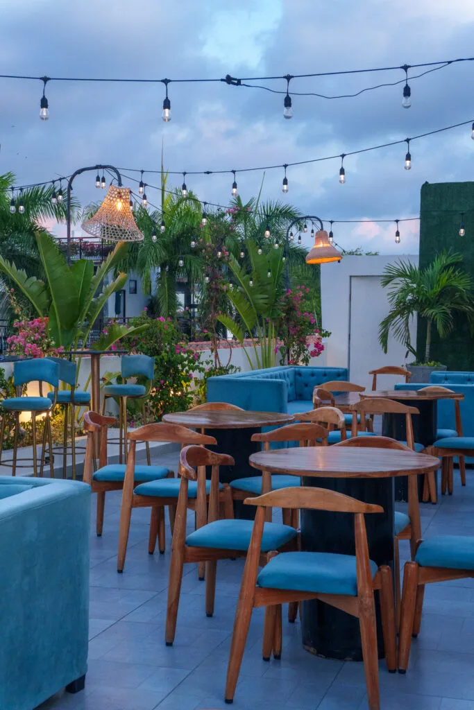 Outdoor rooftop café with wooden tables and blue chairs, surrounded by tropical plants and string lights overhead, creating a cozy and inviting atmosphere at dusk.
