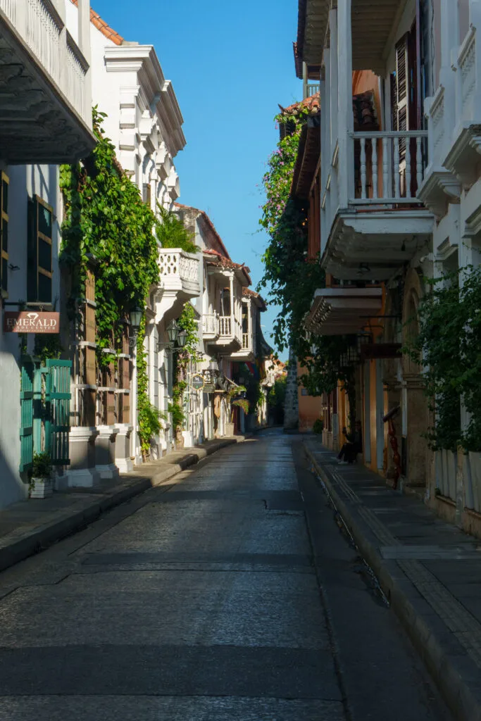 A narrow, empty cobblestone street lined with white colonial-style buildings, balconies, and green plants, under a clear blue sky.