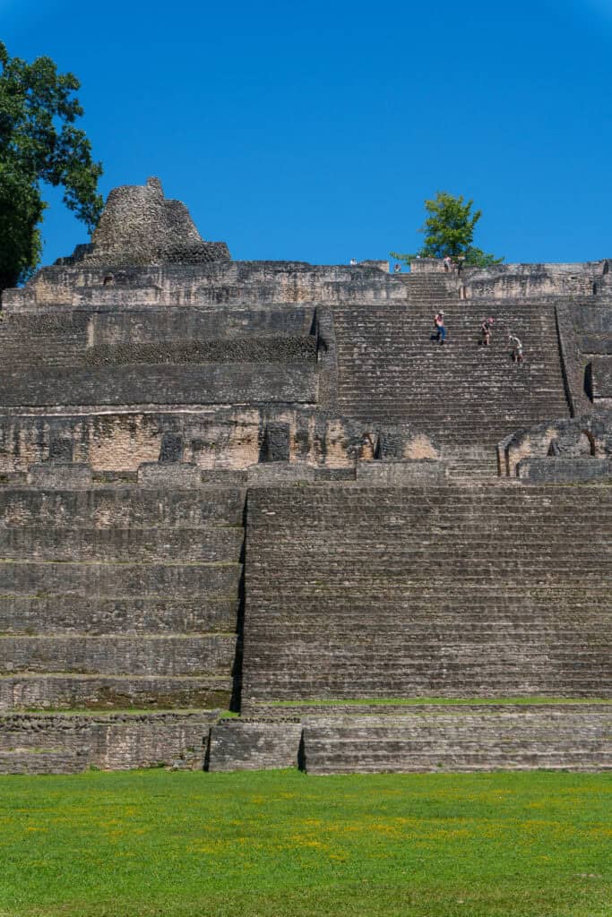 Ancient stone steps of a large Maya pyramid rise against a clear blue sky, with a few people climbing near the top and green grass in the foreground—a striking reminder of the grandeur once found in the ancient cities of Central America during the height of Maya civilization.