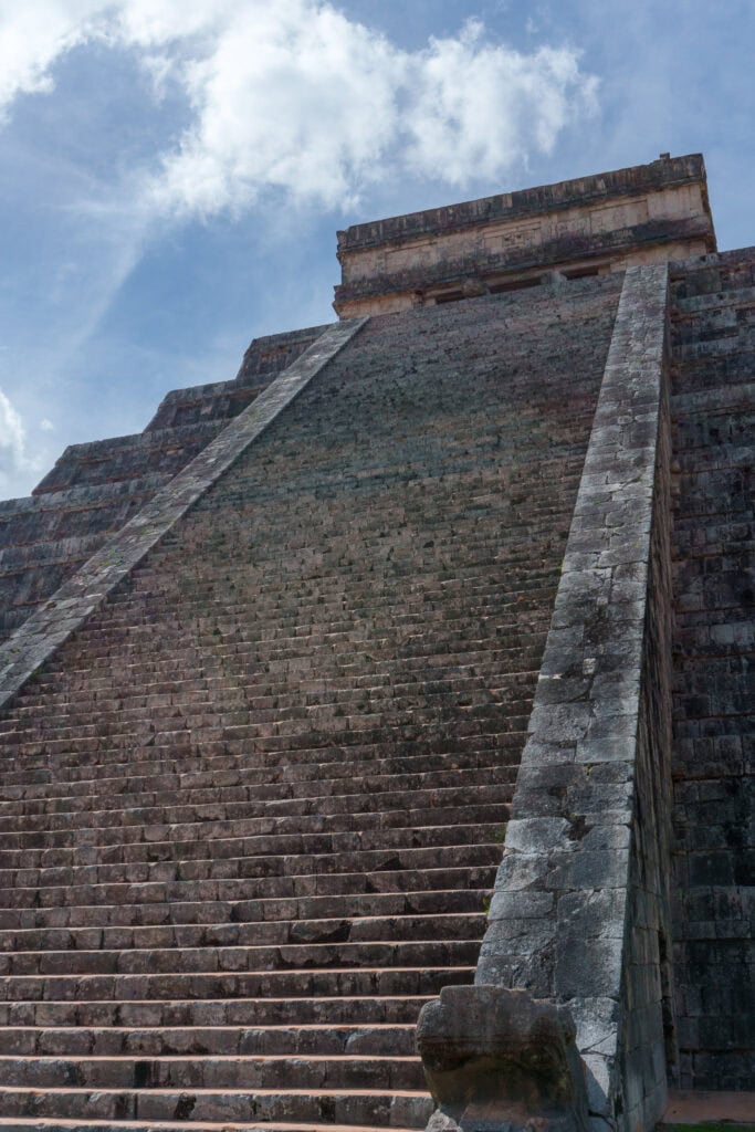 Steep stone steps lead up to the ancient pyramid of Chichen Itza, a renowned Maya site in Mexico, under a blue sky with scattered clouds.