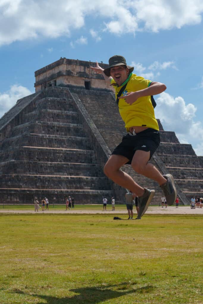 A person in a yellow shirt and hat jumps mid-air in front of the Chichen Itza pyramid, one of the most renowned ancient cities of Central America built by the Maya, on a sunny day, with people walking in the background.