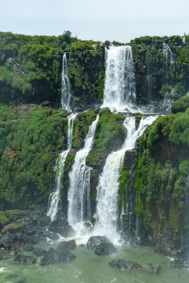 Tall waterfall cascades down mossy, green cliffs into a rocky pool below, surrounded by lush vegetation and mist rising from the water.