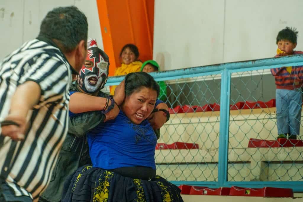 Wrestling in El Alto in La Paz with a cholita fighting a cqtcher with a mask. There is an referee in front and children assisting the spectacle in the background.