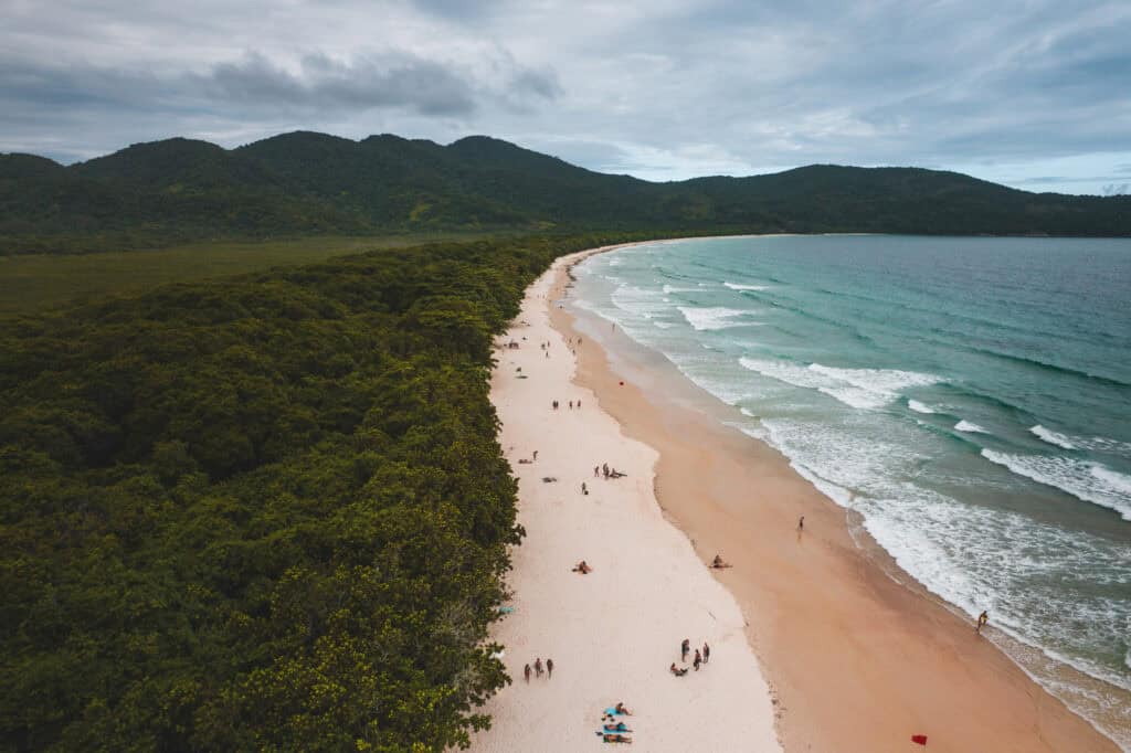 A sandy beach curves along the shoreline with gentle waves, bordered by dense green forest and distant hills under a cloudy sky. People are scattered along the beach, enjoying the scenery, perhaps planning how to get to Ilha Grande from Rio or wondering about things to do in Ilha Grande once they arrive.