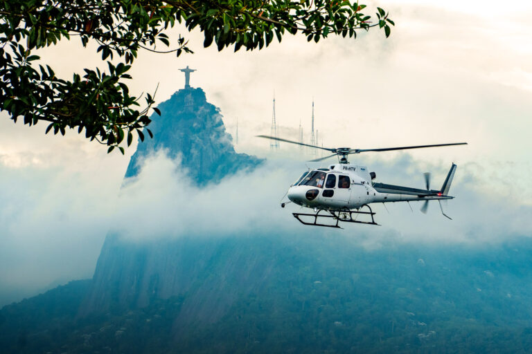 A white helicopter flies near a misty mountain with the Christ the Redeemer statue in the background, framed by green tree branches above—a breathtaking moment often experienced during a rio helicopter tour.