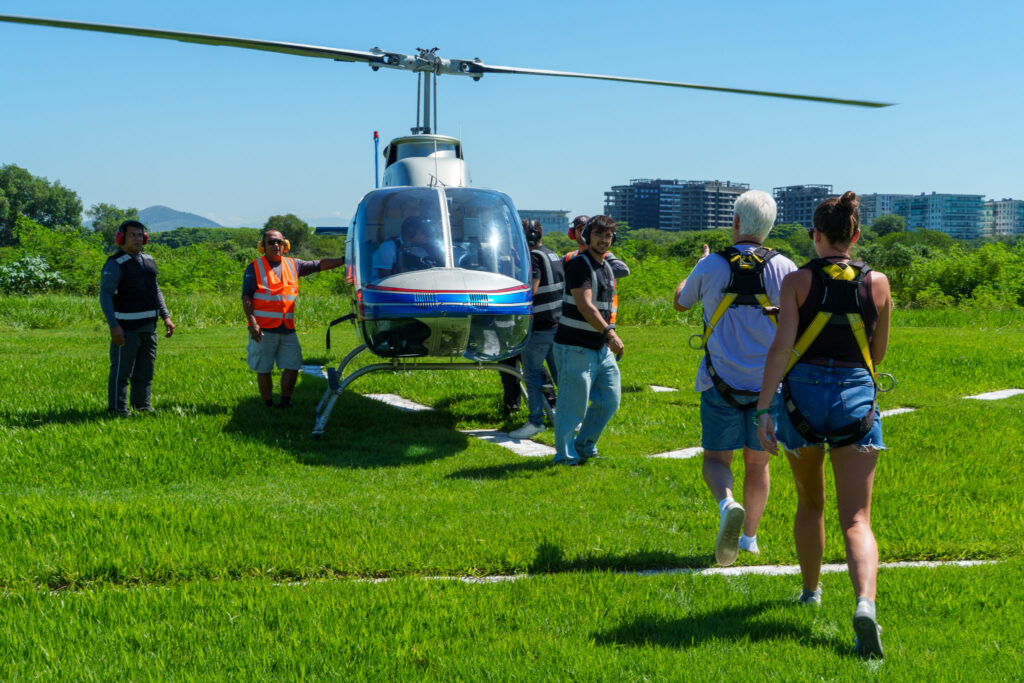 A group of people wearing harnesses walk toward a helicopter on a grassy field, preparing for an unforgettable adventure as part of a helicopter tour Rio experience, while others stand nearby under a clear blue sky.