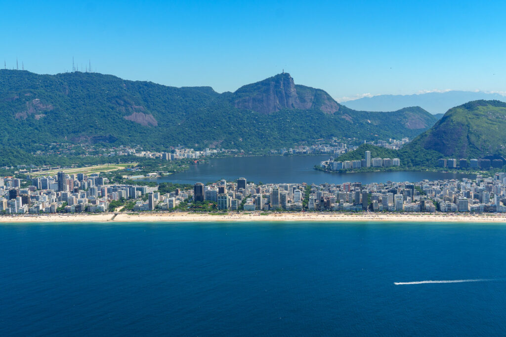 Aerial view of Rio de Janeiro showing a long sandy beach, blue ocean, city buildings, a lagoon, and green mountains under a clear blue sky—the breathtaking scenery you can experience firsthand on a helicopter tour Rio de Janeiro offers. For those curious about the helicopter tour Rio de Janeiro price, it’s an unforgettable way to see the city from above.