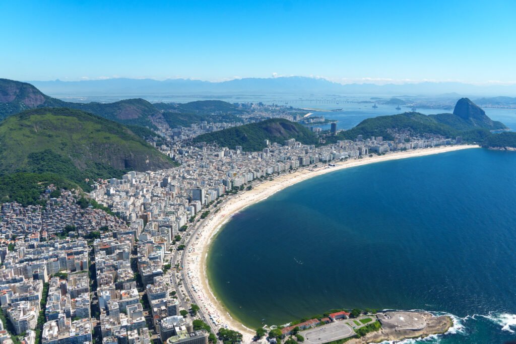 Aerial view of Rio de Janeiro’s Copacabana Beach, with high-rise buildings, green hills, and Sugarloaf Mountain in the background under a clear blue sky—a breathtaking scene best experienced on a helicopter tour of Rio de Janeiro.