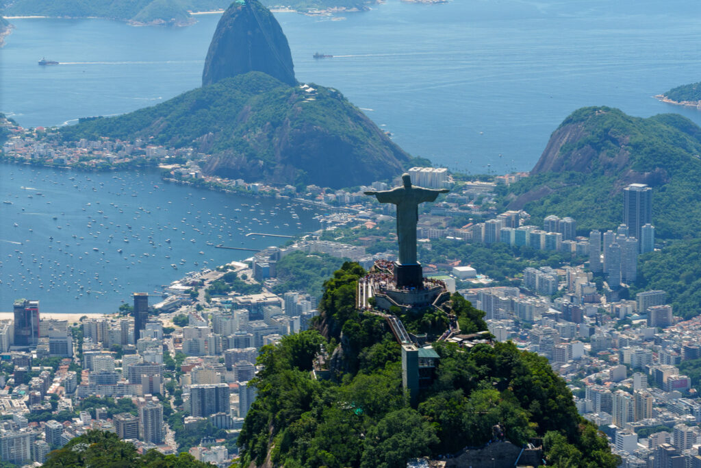 Aerial view of Rio de Janeiro with the Christ the Redeemer statue in the foreground, Sugarloaf Mountain, boats in the bay, and city buildings surrounded by green hills in the background—a scene best experienced on a thrilling rio helicopter tour. If you’re curious about a helicopter tour Rio de Janeiro price, this unforgettable perspective makes it well worth exploring.