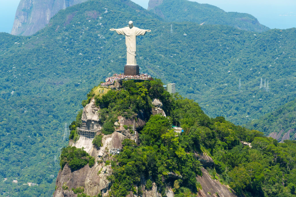 The Christ the Redeemer statue stands atop a lush, green mountain in Rio de Janeiro, Brazil, with visitors gathered at its base and forested hills in the background—a sight that is especially breathtaking when experienced from above during a helicopter tour Rio de Janeiro.