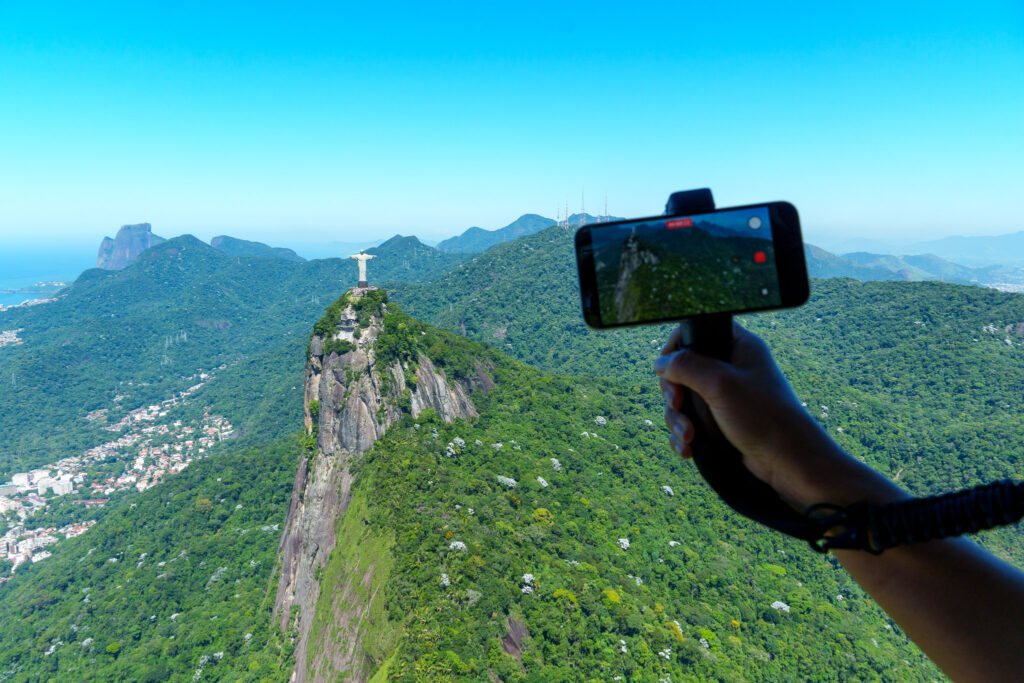 A person films the Christ the Redeemer statue atop a green mountain in Rio de Janeiro with a smartphone, capturing the scenic landscape under a clear blue sky—offering a glimpse into the breathtaking views you might experience during a helicopter tour Rio de Janeiro.