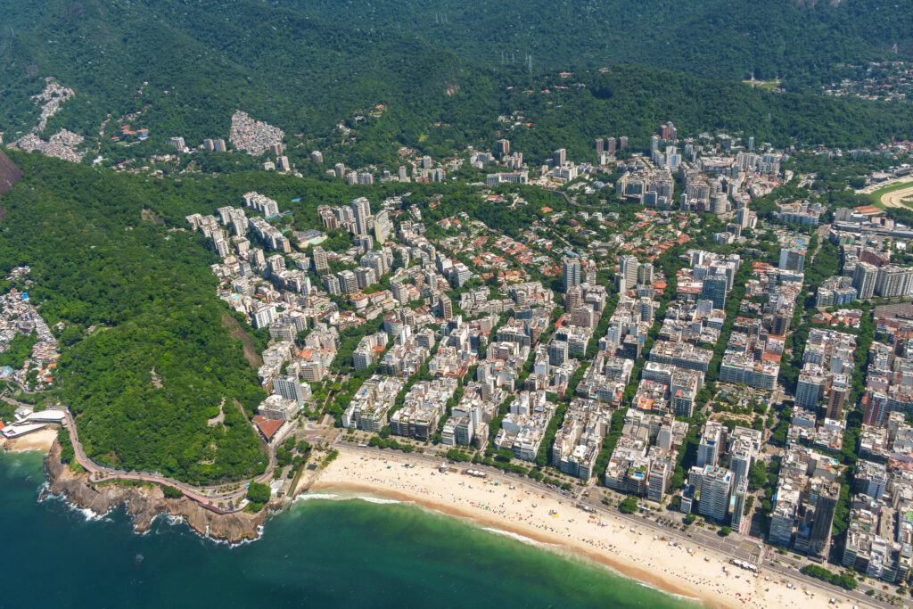 Aerial view of a city with tall buildings, a sandy beach along the shoreline, green forested hills, and blue ocean water. The beach has people and the cityscape extends toward the mountains—capturing the stunning perspective you can experience on a helicopter tour Rio de Janeiro.
