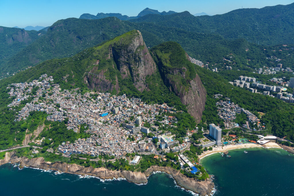 Aerial view of a densely packed coastal city with white buildings, green mountains in the background, and blue ocean waters in the foreground. Sandy beaches curve along the shoreline, showcasing the stunning scenery you can experience on a helicopter tour Rio de Janeiro offers.