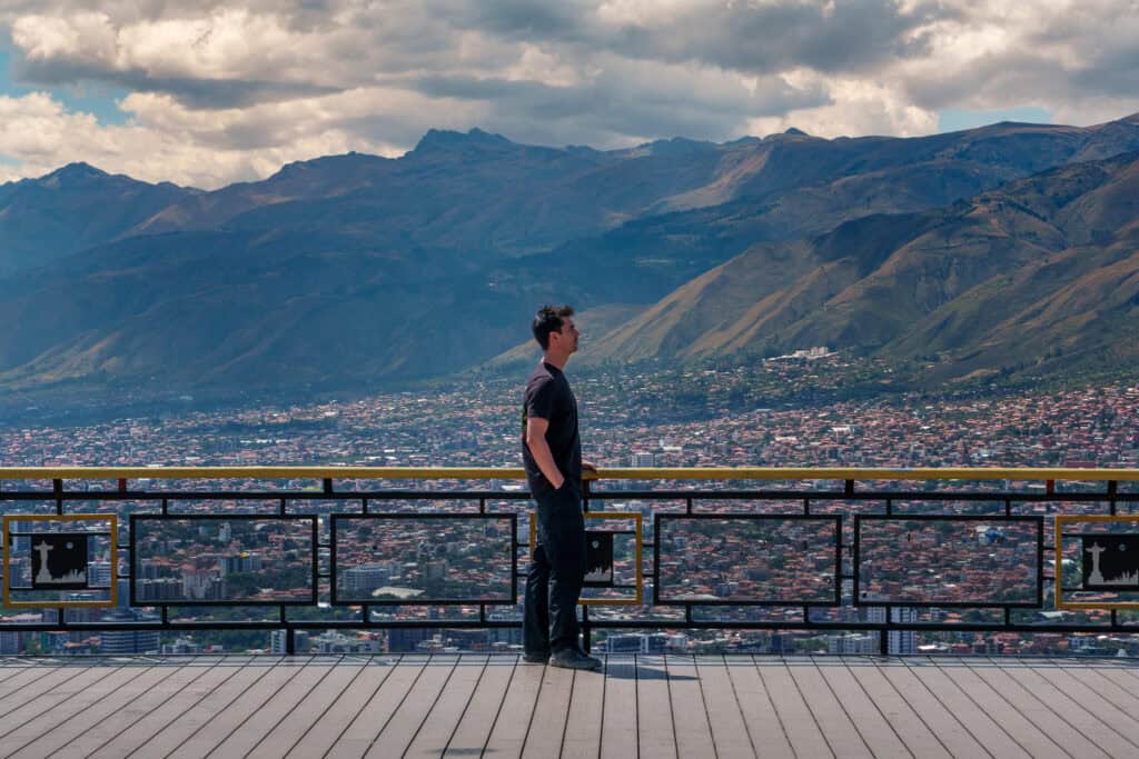 Man in black clothes stands on a platform with decorative railing, overlooking the city of Cochabamba and distant mountains under a cloudy sky, capturing the essence of typical Cochabamba weather.