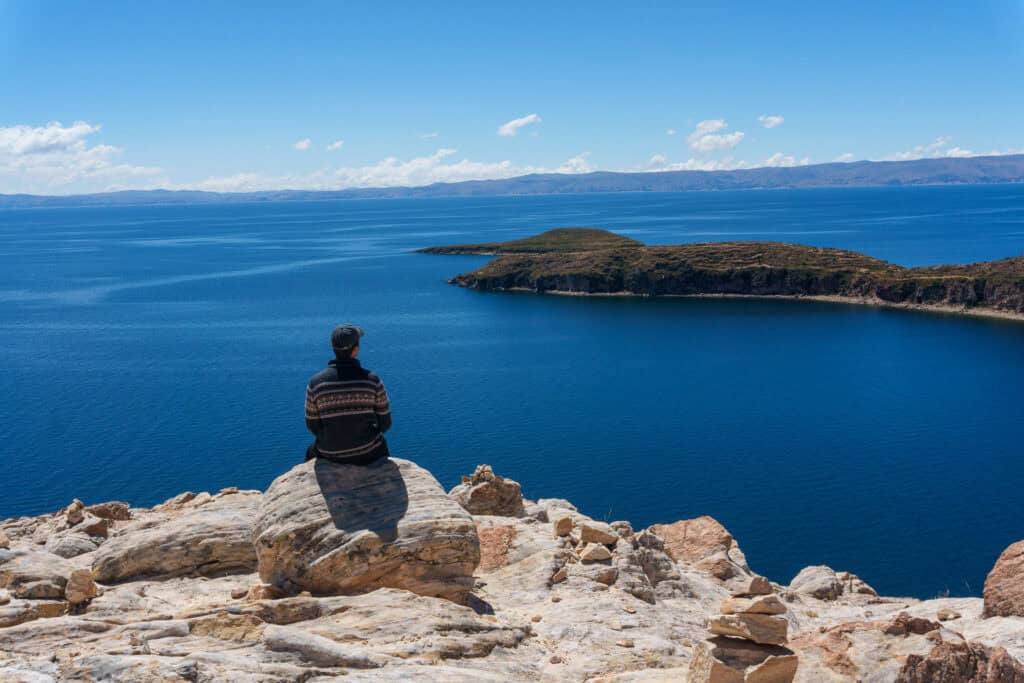 A person sits on a rocky cliff overlooking the vast blue waters of Copacabana Lake Titicaca, with a small island and distant mountains under a clear sky.