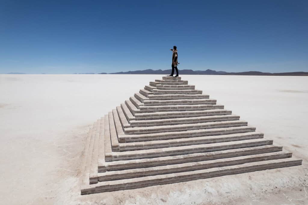 A person stands atop a stepped, pyramid-like structure in the vast, flat expanse of Salar de Uyuni—a dazzling white desert with distant mountains under a clear blue sky.