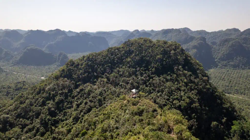 A small structure sits atop a lush, green hill surrounded by dense forest on Cat Ba Island, with layers of rolling, tree-covered mountains visible in the background under a clear sky—a serene scene reminiscent of the tranquil Cat Ba Island beaches in Vietnam.