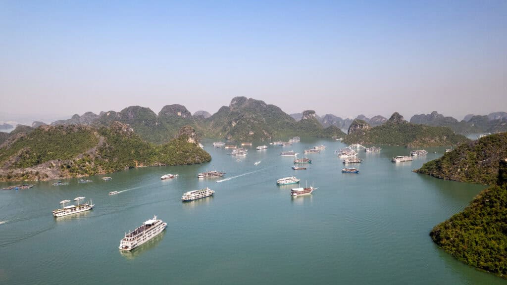 Aerial view of numerous boats sailing in a calm bay surrounded by green islands and rocky hills under a clear sky, capturing the natural beauty and serene atmosphere of Cat Ba Island, Vietnam.