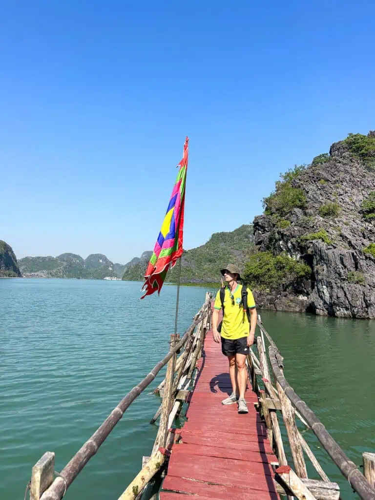 A person in a yellow shirt stands on a narrow wooden bridge over green water beside a colorful flag, with the rocky hills of Cat Ba and a clear blue sky in the background—a picturesque scene often enjoyed by visitors exploring Cat Ba cave or staying at nearby Cat Ba hotels.