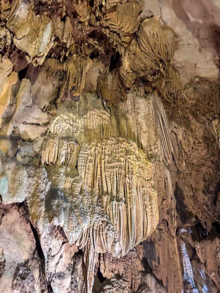 Stalactites and flowstone formations hang from a cave ceiling, displaying textured and layered patterns in shades of brown and tan, illuminated by soft lighting—a scene reminiscent of the stunning limestone caves found on Cat Ba Island in northern Vietnam.