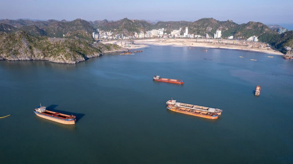 Three large cargo ships float in a calm bay near Cat Ba, a coastal city with hills in the background, under a clear sky.