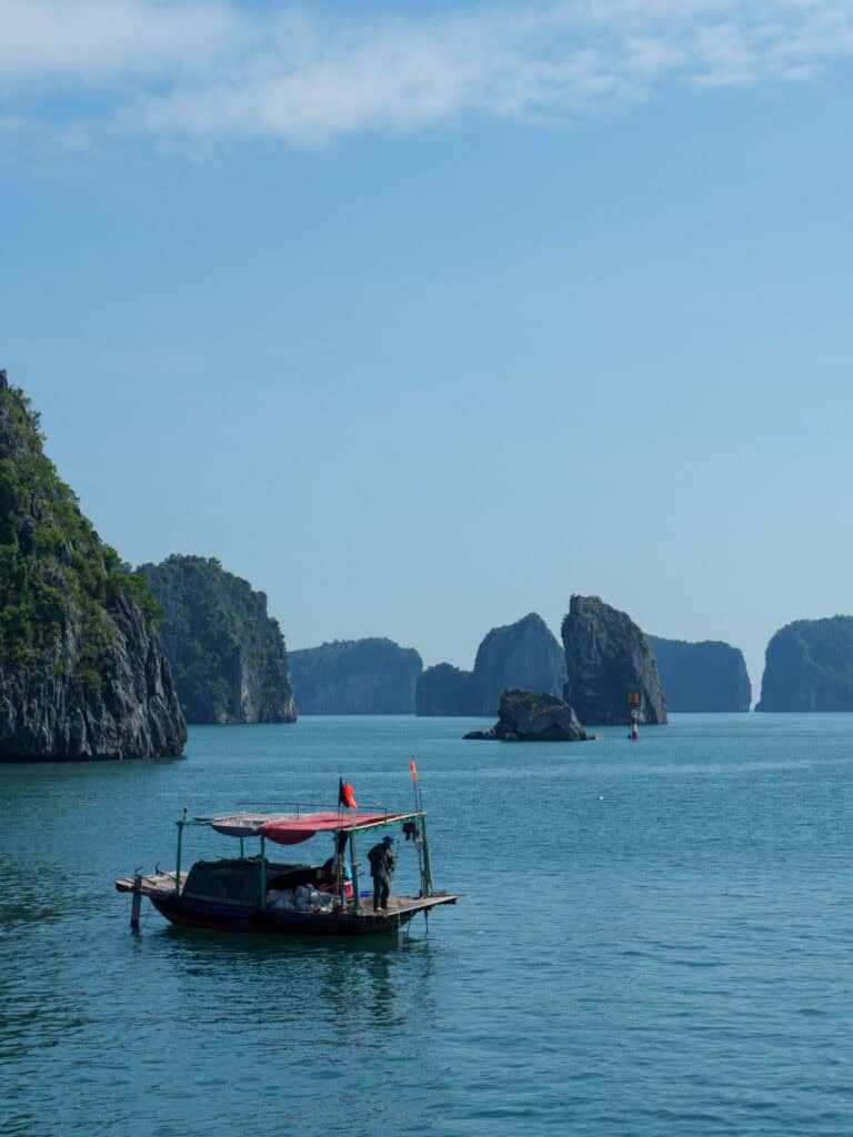 A small boat with a canopy floats on calm blue water, surrounded by the green rocky islands of Cat Ba under a clear sky. Two people are visible standing on the boat.