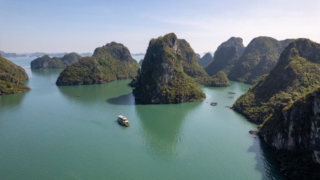 Aerial view of boats sailing among green, rocky islands in calm blue water under a clear sky, with lush vegetation covering the steep cliffs of Cat Ba Island, home to a variety of charming Cat Ba hotels nestled along its picturesque shores.