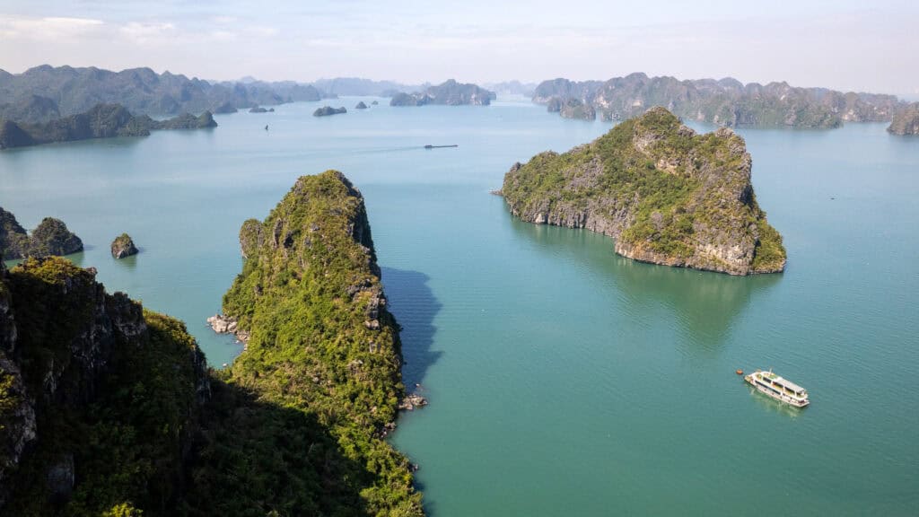 Aerial view of a boat sailing among green, rocky islands in calm blue waters near Cat Ba, Vietnam, with distant islets and a hazy sky in the background.
