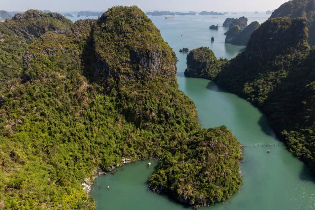 Aerial view of Ha Long Bay, Vietnam, showing lush green islands, calm turquoise water, and scattered boats under a clear sky, with the stunning Cat Ba Island and its famous beaches visible in the distance.