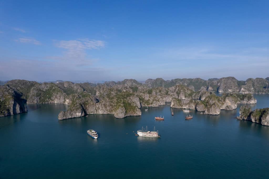 Aerial view of several boats sailing among limestone islands in calm blue water under a clear sky, with rocky green islets scattered across the horizon—an iconic scene near Cat Ba Island, home to some of the most beautiful beaches in Vietnam.