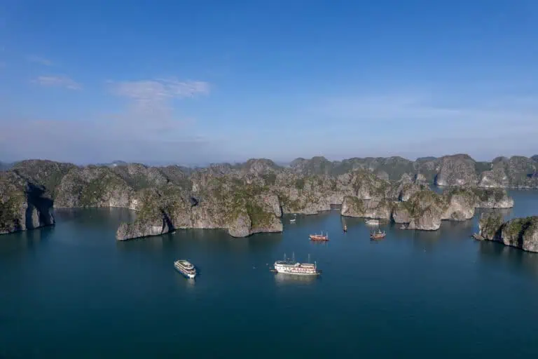 Aerial view of several boats sailing among limestone islands in calm blue water under a clear sky, with rocky green islets scattered across the horizon—an iconic scene near Cat Ba Island, home to some of the most beautiful beaches in Vietnam.