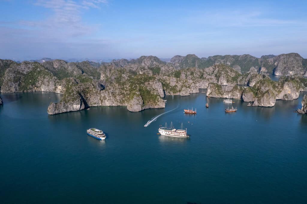 Aerial view of boats sailing among limestone islands and blue water in Lan Ha Bay, Vietnam, with green hills and cliffs in the background under a clear sky.
