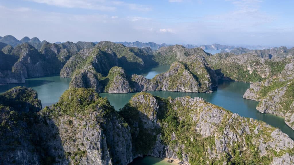 Aerial view of green, rocky islands surrounded by calm blue water under a partly cloudy sky, with distant mountains in the background—this breathtaking landscape is characteristic of Lan Ha Bay Vietnam.