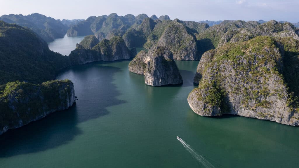 Aerial view of a boat leaving a white trail in green water, surrounded by lush, rocky islands and dramatic limestone cliffs under a clear sky—a stunning scene that perfectly captures the beauty of Lan Ha Bay Vietnam.