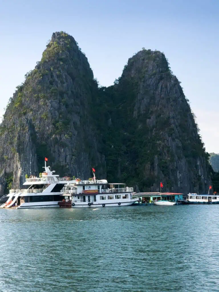 Tour boats with red flags dock by towering limestone cliffs covered in greenery, rising from the calm blue-green water under a clear sky—a picturesque scene often found near Cat Ba Island beaches, highlighting the natural beauty of Cat Ba Vietnam.