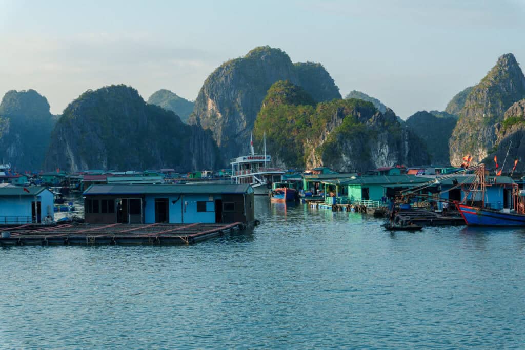 Floating houses and boats on calm water, surrounded by green limestone hills under a partly cloudy sky, create a tranquil scene reminiscent of Cat Ba Island in Vietnam.