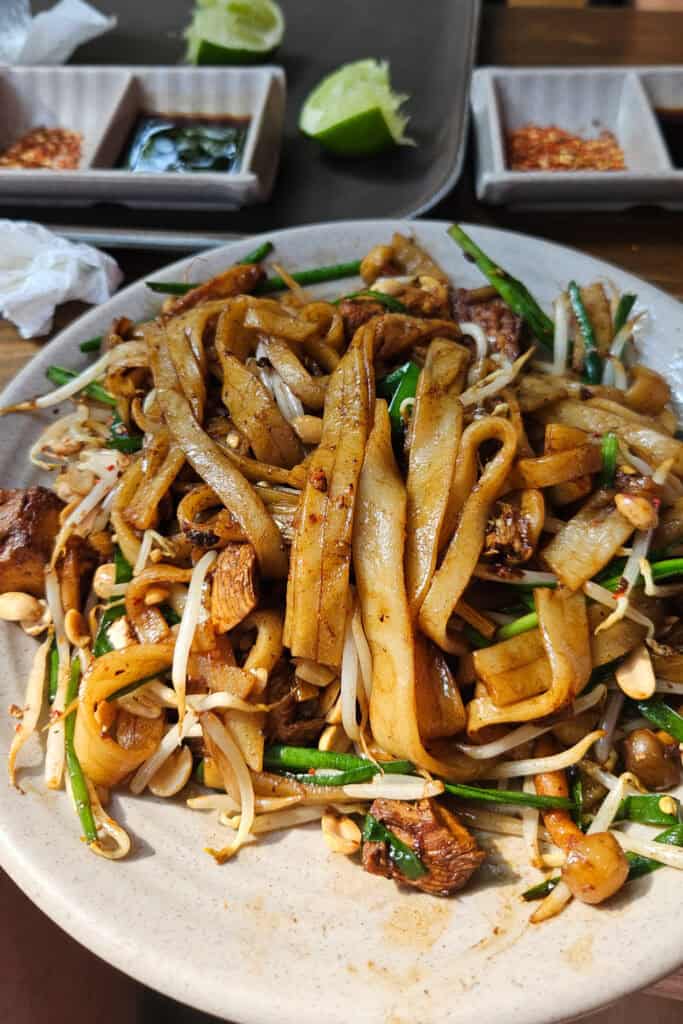 A plate of stir-fried noodles with tofu, bean sprouts, chives, peanuts, and sauce sits on a table—a delicious highlight often recommended in any Saigon travel guide. Lime wedges, chili flakes, and dipping sauce are in the background, capturing the vibrant flavors you’d discover on your culinary journey through Ho Chi Minh City.