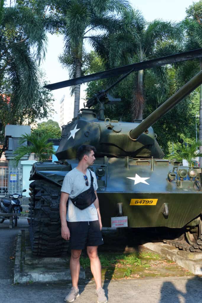 A man in a gray shirt and black shorts stands in front of a green military tank with white stars, surrounded by palm trees and buildings—a scene straight out of any Ho Chi Minh City travel guide.