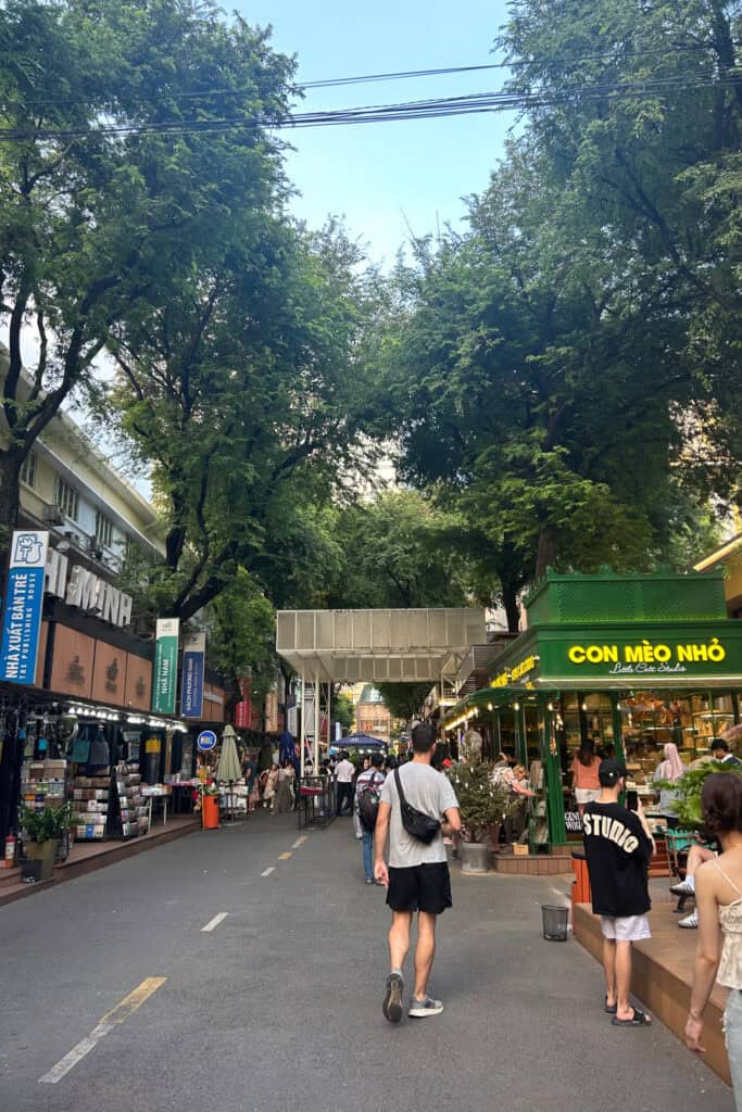 People walk along a tree-lined street with bookshops and stalls on both sides—an attraction in Saigon often highlighted in any good Saigon travel guide. Signs and storefronts are visible, and the sky is clear above.