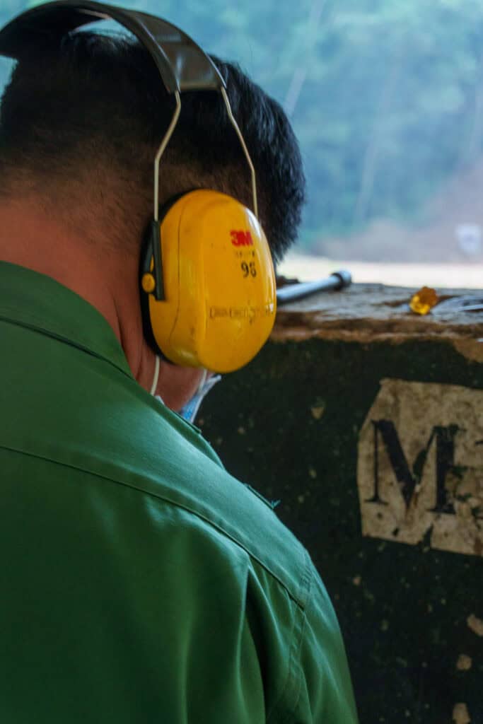 A person in a green shirt wearing yellow protective earmuffs stands next to a weathered wall with a partial letter M visible, evoking the historic atmosphere of the Cu Chi tunnels. The background is blurred trees, reminiscent of the lush landscapes surrounding Cu Chi, Vietnam.