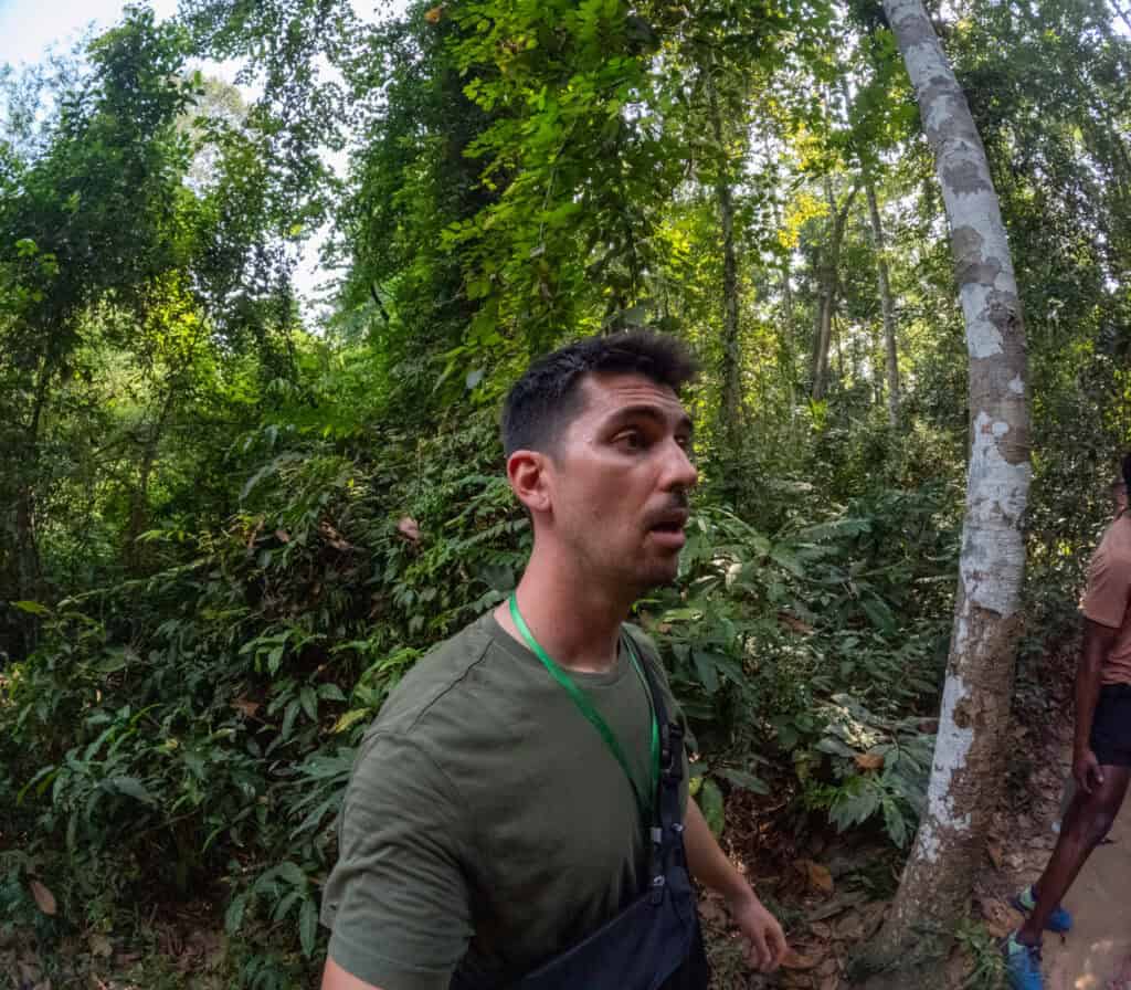 A person with short dark hair wearing a green shirt and a crossbody bag walks through a lush, green forest in Cu Chi, Vietnam. Another person is partially visible to the right. Dense foliage surrounds the scene.