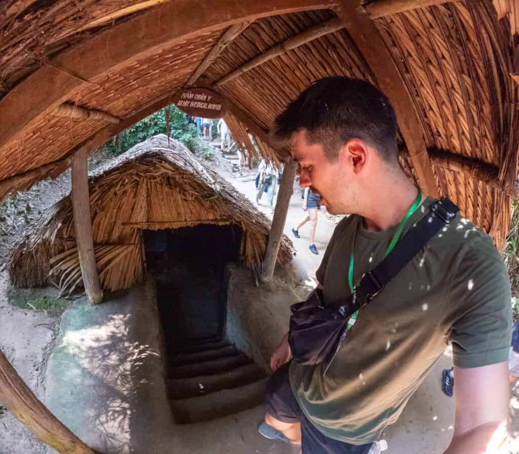 A man stands at the entrance of a narrow, thatched-roof tunnel with stairs leading down, likely at a historical site such as the Cu Chi Tunnels in Vietnam, surrounded by wooden and straw structures.