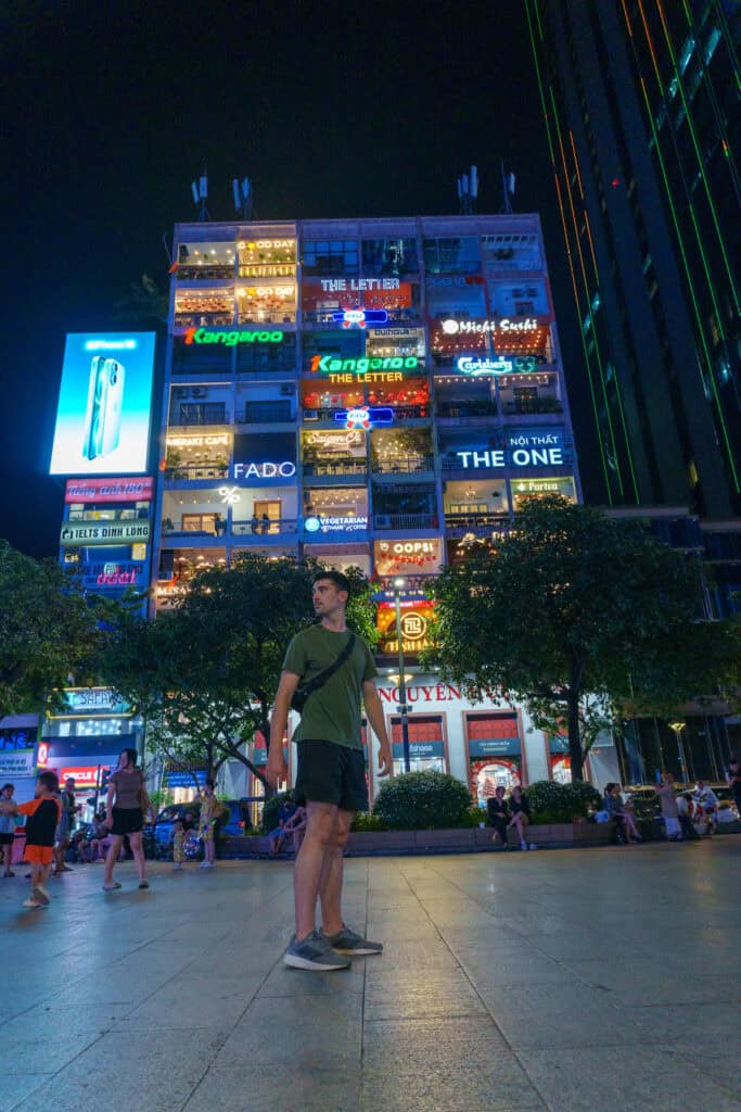 A man stands in a plaza at night in front of a brightly lit building covered in colorful signs and advertisements, evocative of scenes from the Ben Tan Market Saigon section of any Ho Chi Minh City travel guide. People sit and walk nearby, while trees frame the bustling scene.