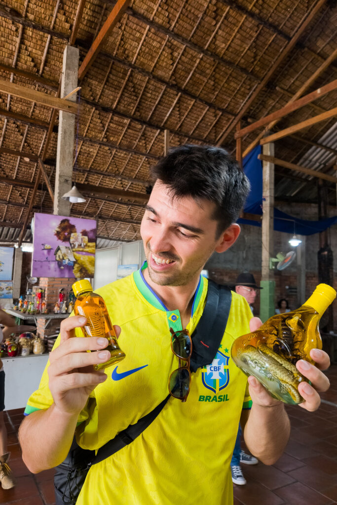 A smiling man in a yellow Brazil jersey holds two bottles of yellow liquid, one containing a snake. He stands inside a rustic market with a thatched ceiling and wooden beams—a scene you might encounter on an authentic Vietnam Cambodia tour as you journey through vibrant local markets along the route from Vietnam to Cambodia.
