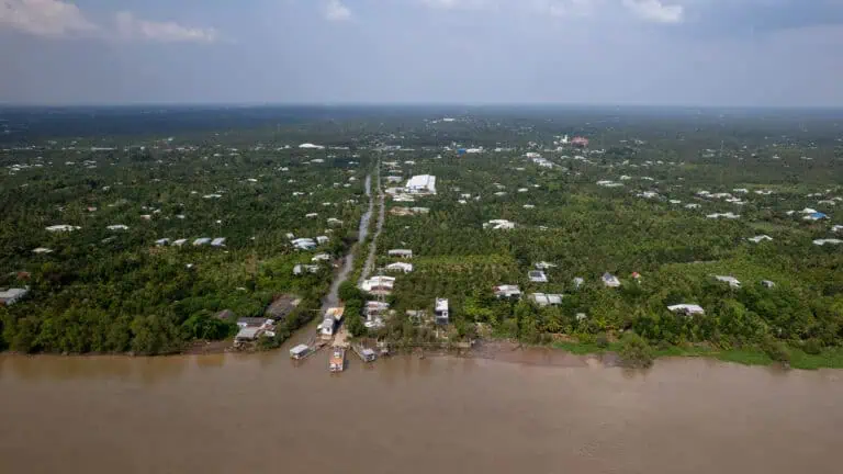 Aerial view of a river with muddy water bordered by green vegetation and scattered houses, a road extends from the river through a lush rural landscape under a cloudy sky—capturing scenes reminiscent of the picturesque routes you might experience on a Vietnam to Cambodia tour, such as traveling from Ho Chi Minh City to Phnom Penh.