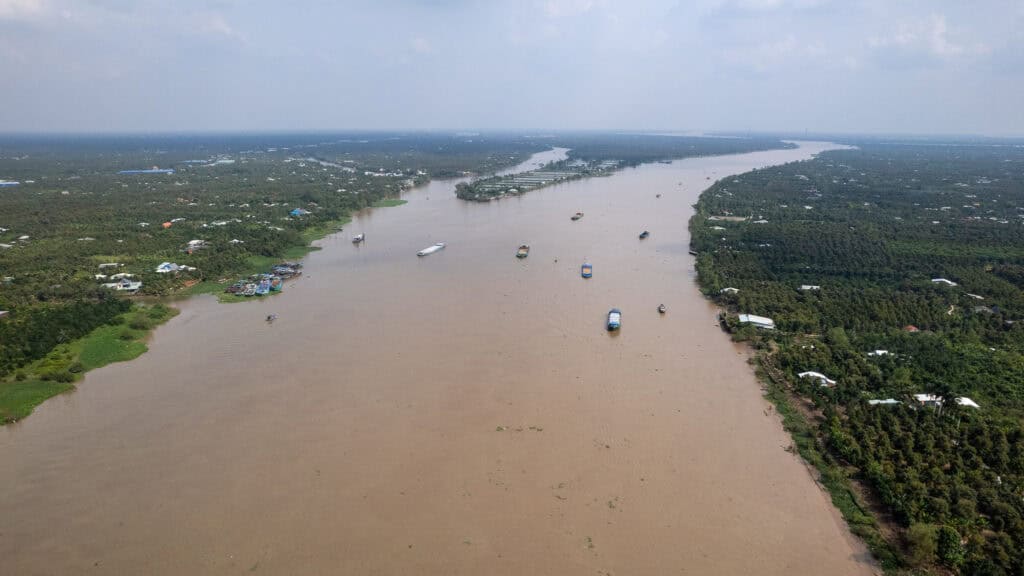 A wide, muddy river flows through a lush green landscape with scattered houses, evoking the scenic journey from Ho Chi Minh to Phnom Penh. Several boats travel along the river under a cloudy sky, capturing the essence of a classic Vietnam Cambodia tour.
