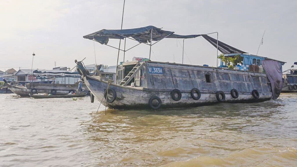 A weathered wooden boat with a canopy floats on murky brown water near a riverside village; smaller boats and buildings are visible in the background, evoking the scenic journey travelers experience on a Vietnam to Cambodia tour.
