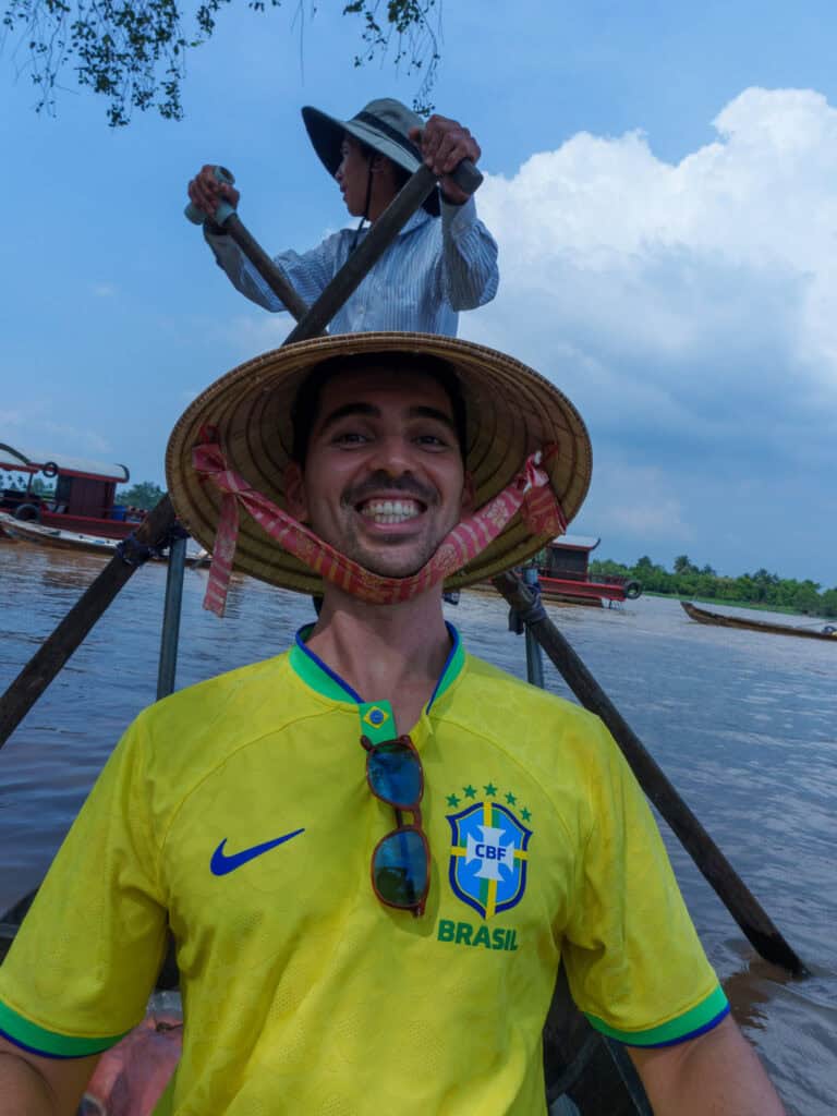 A smiling man in a yellow Brazil jersey and straw hat poses on a boat, with another person rowing behind him on a river under a partly cloudy sky—a perfect scene from a Vietnam to Cambodia tour.