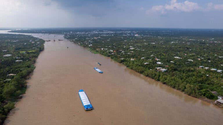 A wide brown river, often traveled on a Vietnam Cambodia tour from Ho Chi Minh City to Phnom Penh, flows past a lush green landscape dotted with houses and several blue-roofed barges under a cloudy sky.